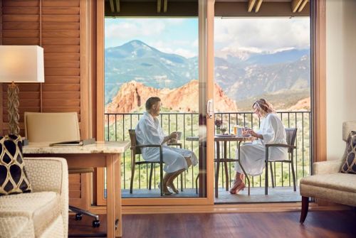Two people in bathrobes sit on a balcony, enjoying breakfast with a scenic mountain view in the background.