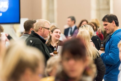 A group of people socializing at an event, some holding drinks, with a blurred background of others mingling.