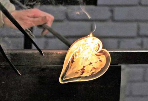A glassblower shapes a glowing heart-shaped glass piece, with smoke rising in a workshop setting.