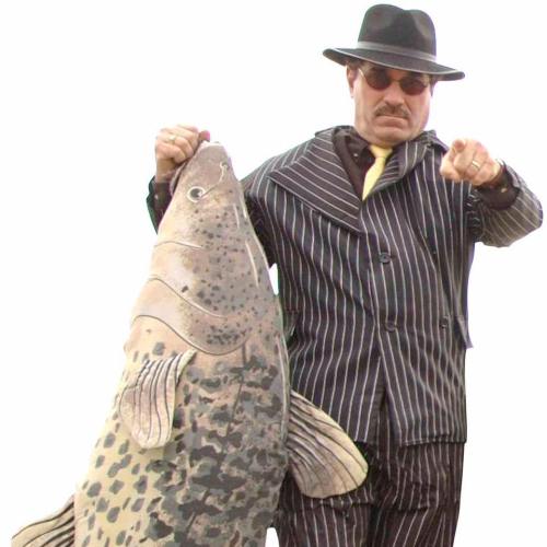 A man in a striped suit and hat holds a large fish, pointing confidently at the camera.