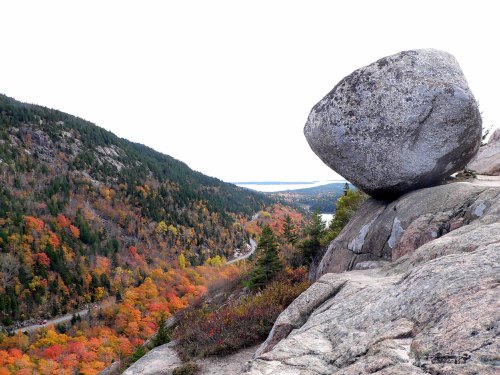 A large boulder perched on a rocky ledge overlooking a colorful autumn landscape with trees and a winding road.