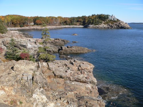 Scenic coastal view with rocky shore, calm blue water, and autumn foliage on trees in the background.
