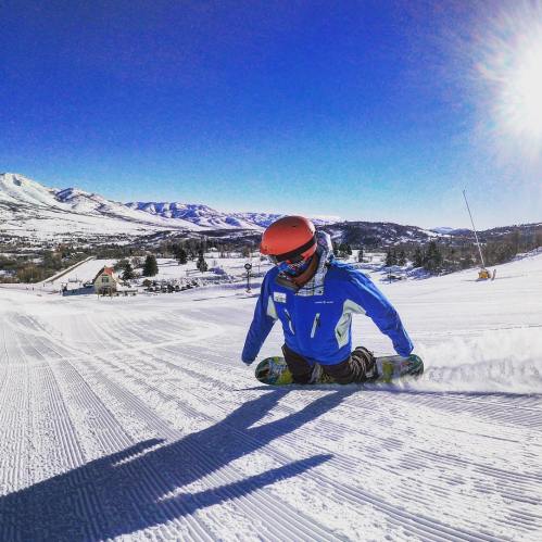 A snowboarder in a blue jacket carves through fresh snow on a sunny mountain slope.
