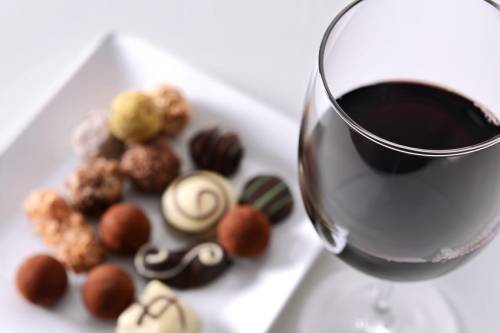 A glass of red wine beside a plate of assorted chocolates and truffles on a white background.