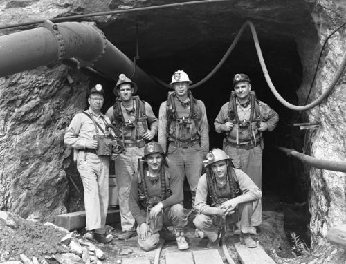 A group of six miners in helmets and gear, posing at the entrance of a mine, with equipment and pipes in the background.