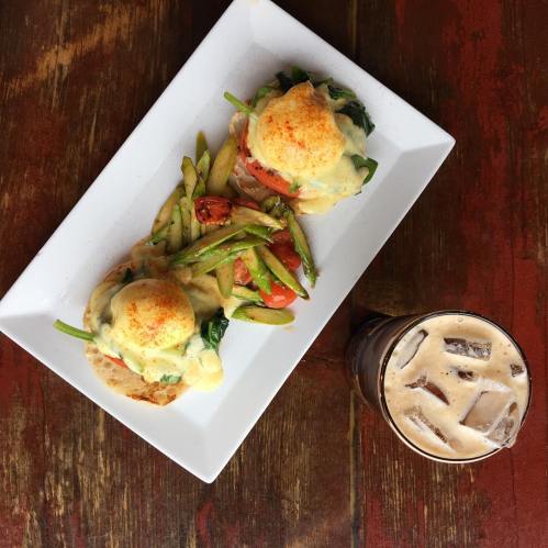 A plate of eggs Benedict with spinach and asparagus, served with a glass of iced coffee on a rustic wooden table.