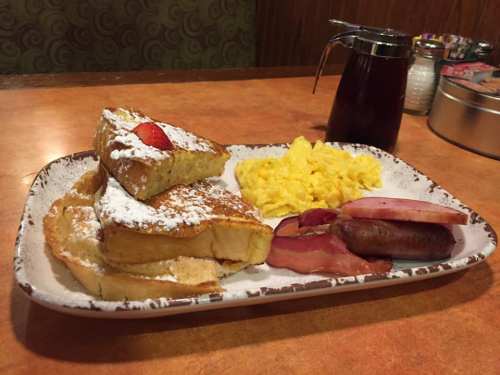 A plate of French toast with powdered sugar, scrambled eggs, bacon, sausage, and a syrup bottle on the side.