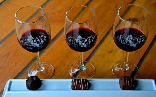 Three wine glasses filled with red wine, accompanied by a plate of chocolate truffles on a wooden table.