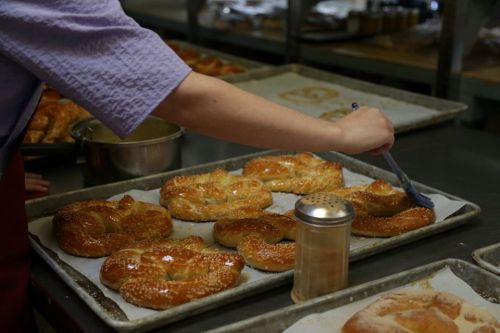 A hand brushes glaze onto freshly baked pretzels on a tray, with a shaker of seasoning nearby.