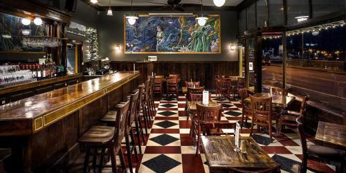 A cozy bar interior with wooden furniture, a patterned floor, and a colorful mural on the wall.