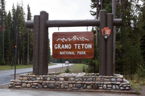 Sign for Grand Teton National Park, featuring wooden structure and stone base, surrounded by trees and a road.