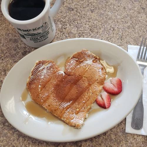 A heart-shaped pancake drizzled with syrup, accompanied by strawberries and a cup of coffee on a table.