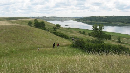 A scenic landscape featuring rolling hills, lush greenery, and a calm river under a cloudy sky, with people walking along a path.