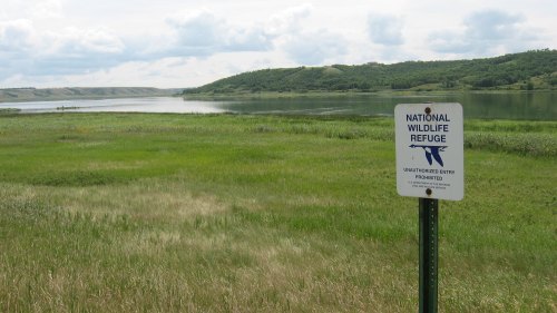 A sign for a National Wildlife Refuge stands in a grassy area near a calm lake and rolling hills under a cloudy sky.