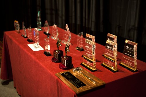 A table displaying various awards and trophies, including glass plaques and wooden honors, on a red tablecloth.