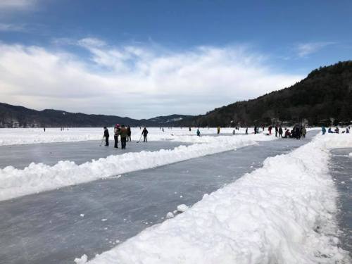 People ice skating on a frozen lake, with snow-covered banks and mountains in the background under a blue sky.