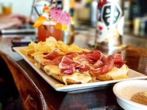 A plate of melted cheese topped with sliced meats, served with chips and a colorful cocktail in the background.