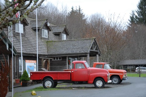 Two vintage red trucks parked in front of a rustic wooden building surrounded by trees.