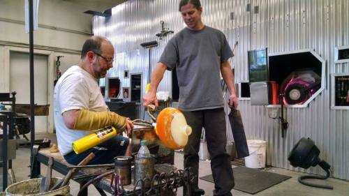Two glassblowers work together in a studio, shaping molten glass with tools and a blowpipe.