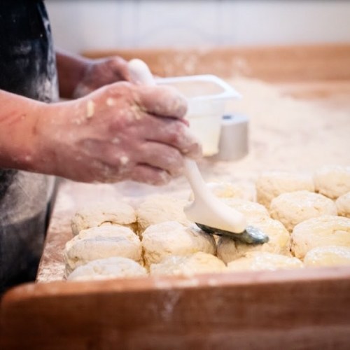 A person brushes dough with a pastry brush on a floured surface, preparing biscuits for baking.