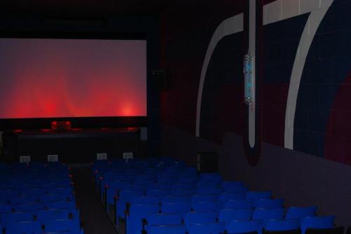Empty movie theater with blue seats and a dimly lit screen, featuring red and dark walls.