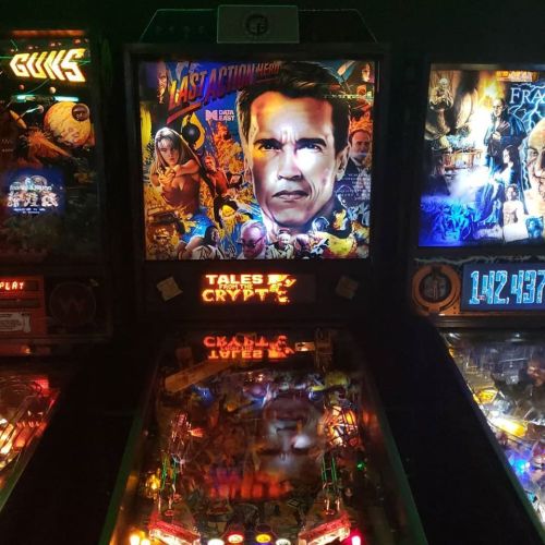 A brightly lit pinball machine featuring "Last Action Hero" artwork and other classic arcade games in a dark setting.