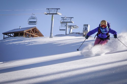 A skier carves through fresh snow on a sunny day, with ski lifts and a lodge in the background.
