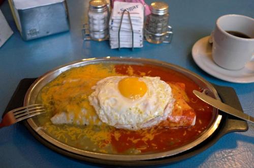 A plate of enchiladas topped with cheese and a fried egg, served with a side of red sauce and a cup of coffee.