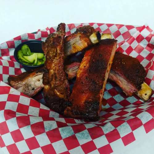 A basket of juicy, smoked ribs with a side of pickles, all served on red and white checkered paper.