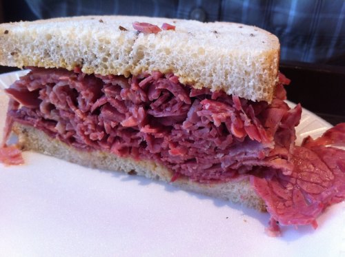 A close-up of a stacked pastrami sandwich on white bread, showcasing layers of tender, pink meat.