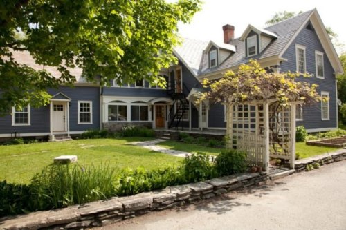 A charming blue house with a white trellis, surrounded by greenery and a stone pathway on a sunny day.