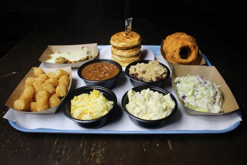 A tray of various comfort foods including tater tots, baked beans, coleslaw, macaroni salad, waffles, and fried chicken.