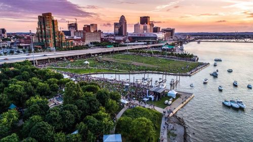 Aerial view of a city skyline at sunset, with a river, boats, and a crowd gathered on the waterfront.