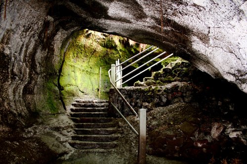 A moss-covered cave entrance with stone steps and metal railings leading into a dimly lit interior.