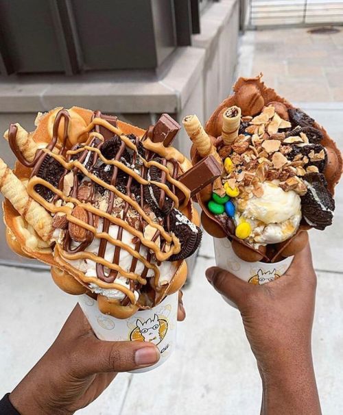 Two hands hold colorful ice cream cones topped with chocolate, cookies, and various candies against an urban backdrop.