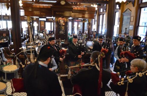 A group of musicians in traditional attire play bagpipes in a lively pub setting.