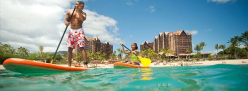 A man and a girl paddleboarding in clear water with a resort in the background and palm trees under a blue sky.