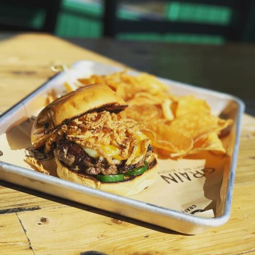 A juicy burger with melted cheese, crispy onions, and pickles, served with a side of potato chips on a wooden table.