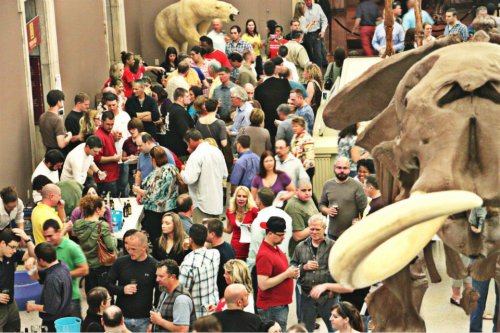 A crowded event in a museum, featuring people socializing and enjoying drinks, with large animal exhibits in the background.