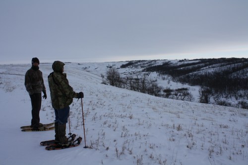 Two people in snowshoes stand on a snowy hillside, overlooking a winter landscape with rolling hills and bare trees.