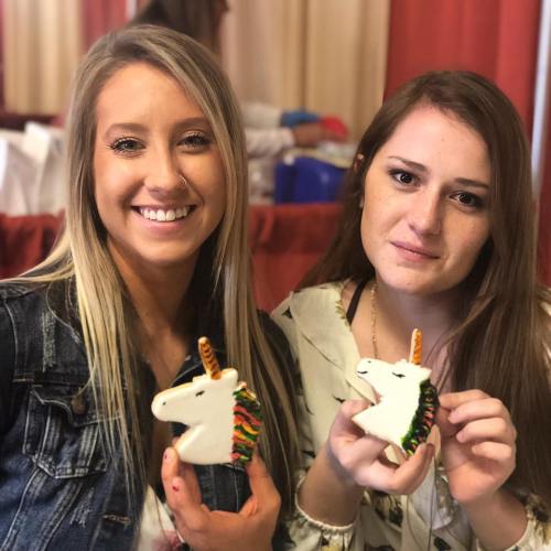 Two smiling women hold colorful unicorn figurines, posing together in a bright indoor setting.