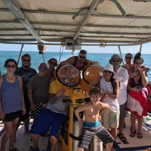 A group of people poses on a boat with a yellow submarine in the background, enjoying a sunny day at sea.