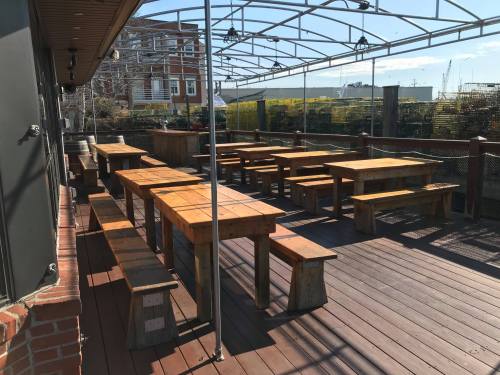 Outdoor dining area with wooden tables and benches under a clear roof, surrounded by greenery and a sunny sky.