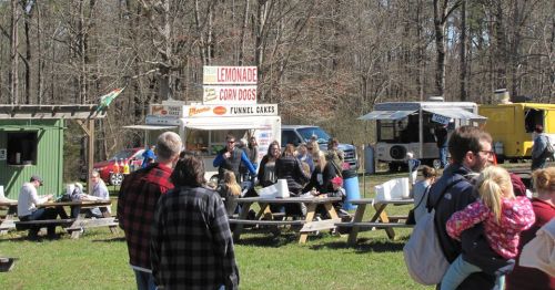 A busy outdoor food festival with food trucks and people enjoying the day in a wooded area.