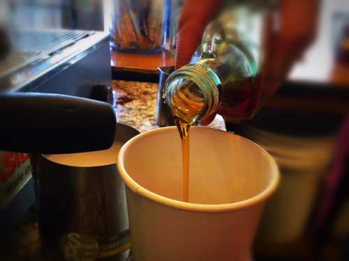 A hand pours syrup from a glass bottle into a white cup, with a coffee machine in the background.