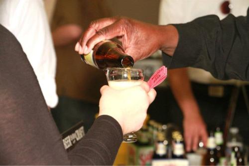 A hand pours a drink into a glass held by another hand, with various bottles in the background.