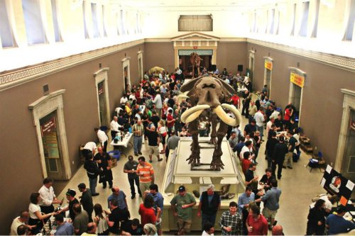 A crowded museum hall featuring a mammoth skeleton and various exhibits, with visitors engaging and exploring.
