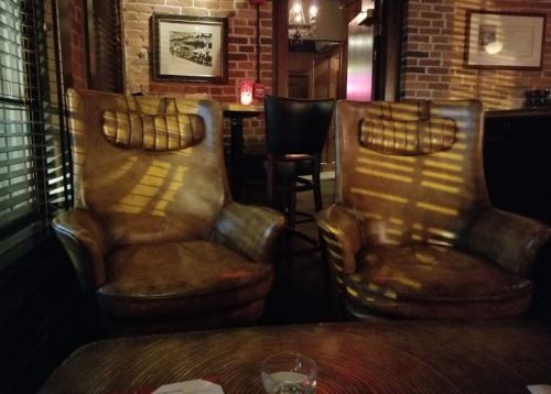 Two vintage leather armchairs with unique headrests, illuminated by soft lighting in a cozy, brick-walled setting.
