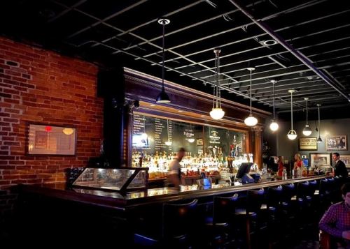 A dimly lit bar with a wooden counter, shelves of liquor, and patrons enjoying drinks in a cozy atmosphere.