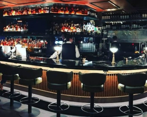 A stylish bar with a curved counter, illuminated bottles, and bartenders in vests serving drinks.